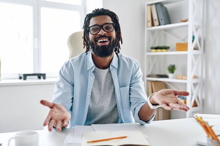 Happy young African man in eyewear keeping arms outstretched and smiling while working indoorsの写真素材
