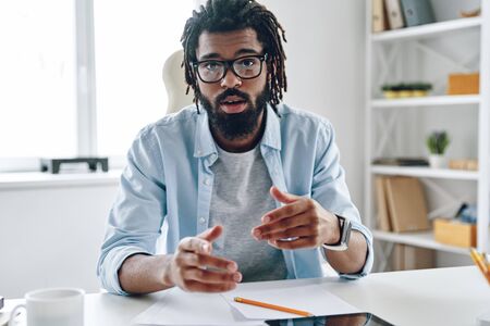 Confident young African man in eyewear telling something and looking at camera while working indoorsの写真素材