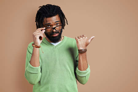 Charming young African man in casual clothing pointing copy space and adjusting eyewear while standing against brown backgroundの写真素材