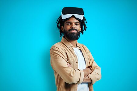 Handsome young African man in virtual reality glasses looking at camera and smiling while standing against blue backgroundの写真素材