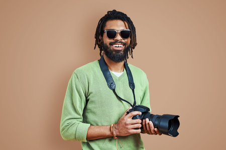 Young African photographer in casual clothing looking at camera and smiling while standing against...
