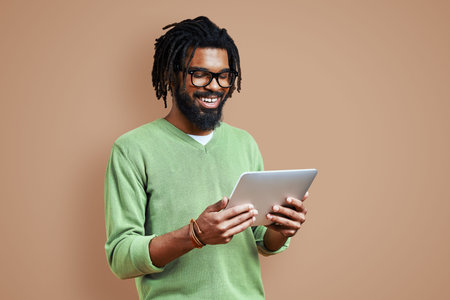 Handsome young African man in smart casual clothing using digital tablet and smiling while standing...