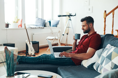 Handsome young man in casual clothing using laptop and smiling while sitting on the sofaの写真素材