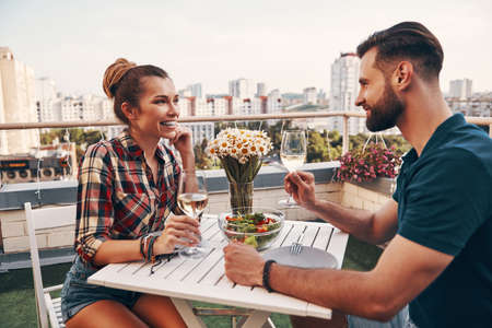Young couple in casual clothing enjoying romantic dinner and smiling while enjoying romantic dinner on the rooftop patio outdoorsの写真素材