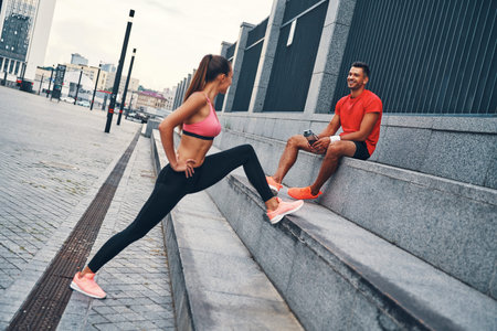 Beautiful young couple in sports clothing warming up and stretching while practicing outdoors on the...