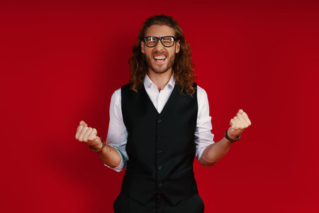 Happy young man in elegant clothing cheering and gesturing while standing against red background