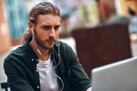 Good looking young man in casual clothing working using computer and headphones while sitting in...