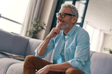 Side view of carefree senior man in casual clothing and eyeglasses holding hand on chin and smiling while resting on the sofa at homeの写真素材