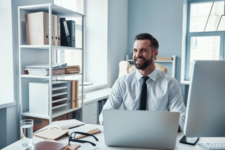 Happy young man in shirt and tie working using computer and smiling while sitting in the office