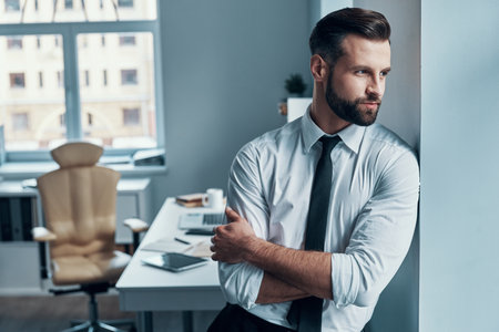 Thoughtful young man in formalwear looking away and keeping arms crossed while standing in officeの写真素材