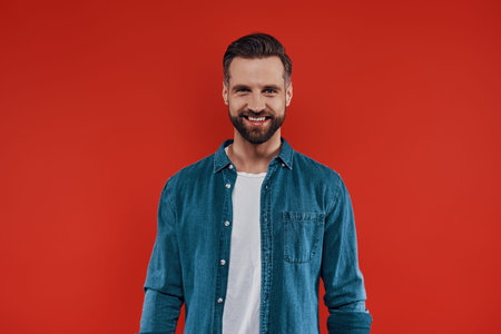 Handsome young man in casual clothing smiling and looking at camera while standing against red...