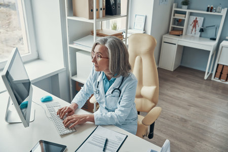 Top view of mature female doctor in white lab coat working on computer while sitting in her office