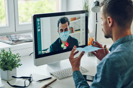 Confident young man in protective face mask talking to collegue by video call while sitting in...