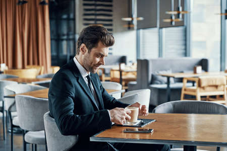 Handsome young businessman in full suit enjoying coffee and smiling while sitting in the restaurantの写真素材