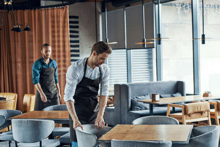 Handsome young men in aprons arranging furniture while preparing restaurant to openingの写真素材
