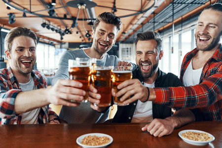 Happy young men in casual clothing toasting each other with beer and laughing while sitting in the...