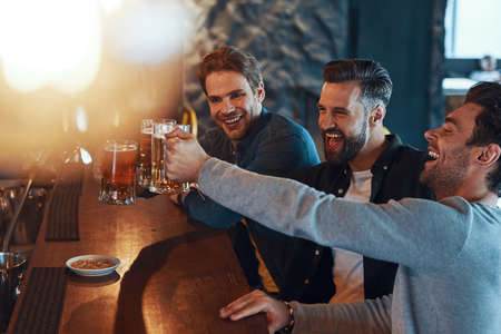 Happy young men in casual clothing toasting each other with beer and laughing while sitting in the pubの写真素材
