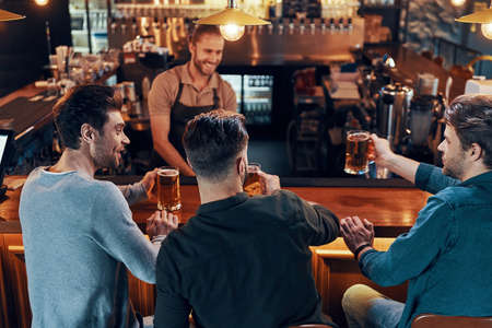 Smiling young men in casual clothing drinking beer while sitting in the pub togetherの写真素材