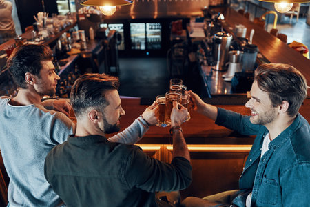 Top view of happy young men in casual clothing toasting each other with beer and smiling while...