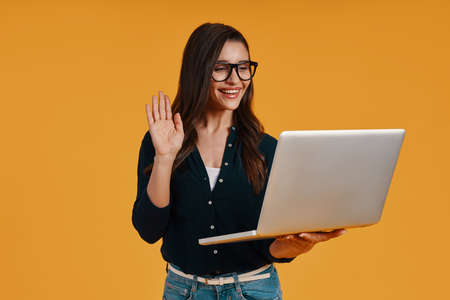 Happy young woman in smart casual clothing using laptop and waving handの写真素材
