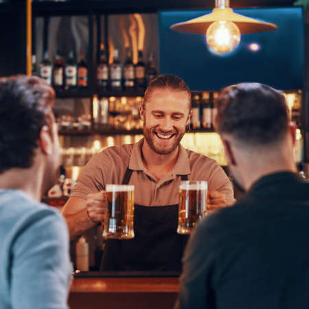 Cheerful bartender serving beer to young men while standing at the bar counter in pubの写真素材