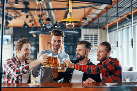 Group of happy young men in casual clothing toasting each other with beer and smilingの写真素材