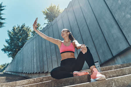 Low angle view of beautiful young woman in sports clothing making selfie by her smart phoneの写真素材