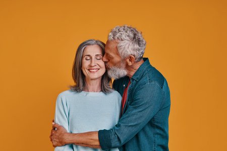 Beautiful senior couple kissing and smiling while standing together against orange backgroundの写真素材