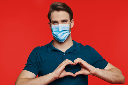 Handsome young man in protective face mask looking at camera and gesturing heart shape