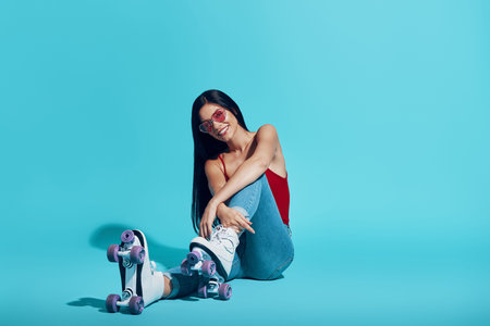 Attractive young woman looking at camera and smiling while wearing roller skates against blue...