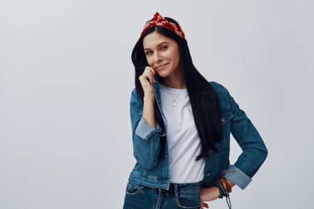 Attractive young woman in bandana looking at camera and smiling while standing against grey...