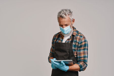 Senior man in apron and protective face mask using digital tablet while standing against gray...