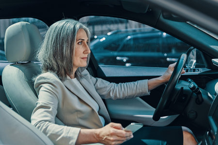 Mature beautiful woman in smart casual wear smiling and using phone while driving car