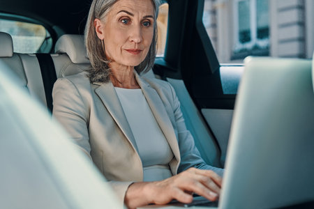 Mature beautiful woman in smart casual wear using laptop while sitting on the back car seat