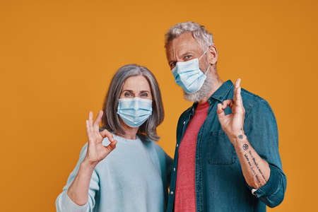Happy senior couple in protective facemasks gesturing while standing together against orange backgroundの写真素材