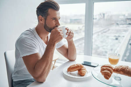 Handsome young man enjoying hot coffee while sitting at the kitchenの写真素材