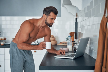 Handsome young shirtless man using laptop while drinking tea at home kitchenの写真素材