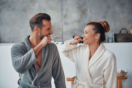 Happy young couple in bathrobes cleaning teeth while doing morning routineの写真素材
