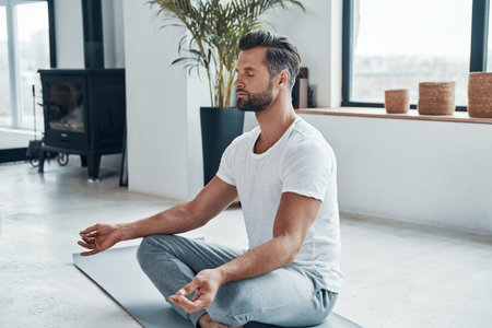 Concentrated young man doing yoga while sitting in lotus position at home