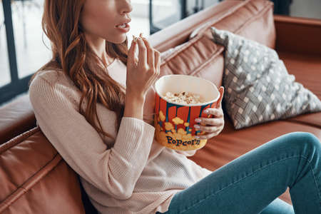Close up of surprised young woman eating popcorn while sitting on the sofaの写真素材