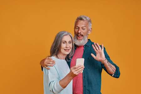 Happy senior couple smiling and using smart phones while standing against orange background