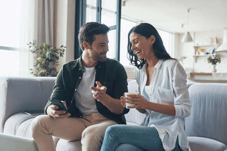 Handsome young showing his girlfriend smart phone and smiling while sitting on the sofa at homeの写真素材