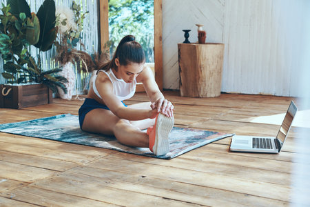 Beautiful young woman in sports clothing stretching on patio with laptop laying near her