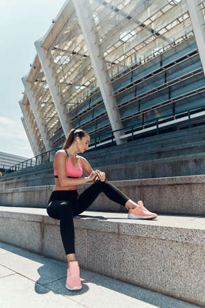 Attractive young woman in sportswear checking her smart watch while sitting outdoorsの写真素材