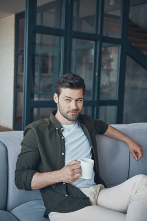 Handsome young man holding cup with drink while sitting on the sofa at home