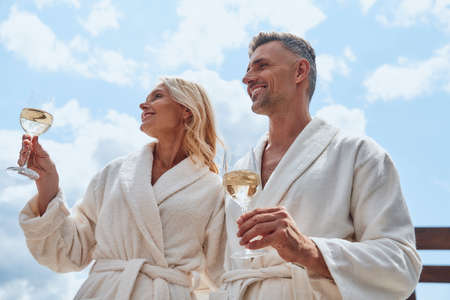 Low angle view of beautiful mature couple in bathrobes enjoying champagne while relaxing outdoorsの写真素材