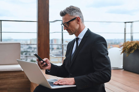 Confident mature man working on laptop and using mobile phone while standing on the rooftop terrace