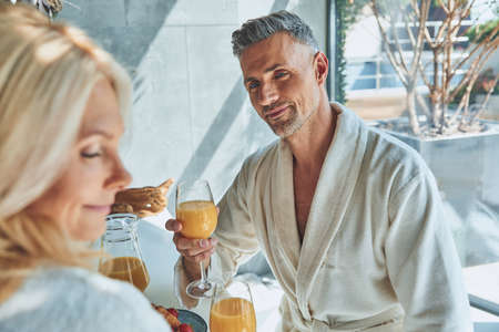 Beautiful mature couple in bathrobes enjoying breakfast together while spending time in the domestic kitchenの写真素材