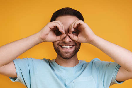Portrait curious young man in casual clothing gesturing eyeglasses and smiling while standing against yellow backgroundの写真素材