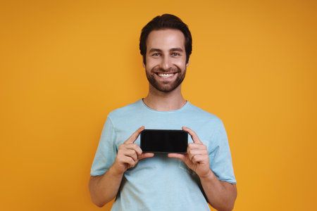 Happy young man in casual clothing showing his smart phone and smilingの写真素材